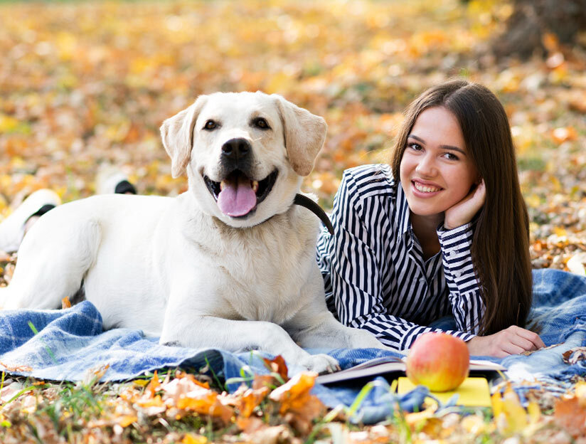 Smiling young woman lying on a blanket next to a white Labrador in a park with autumn leaves, an apple and notebook nearby.